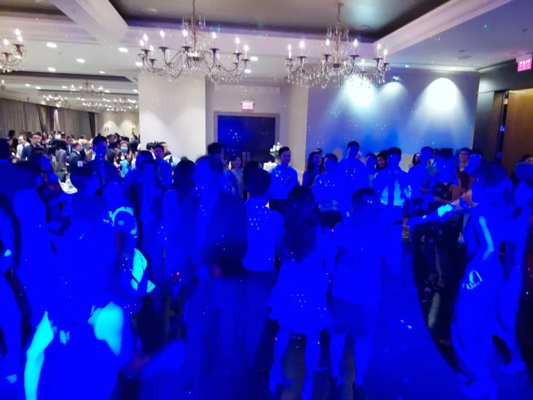 Crowd dancing on a packed ballroom dance floor under blue uplighting and crystal chandeliers