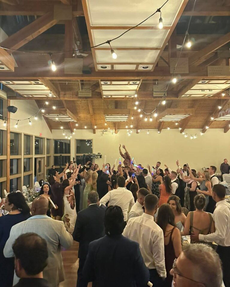 Packed wedding dance floor under wooden beams and string lights with guests' hands in the air